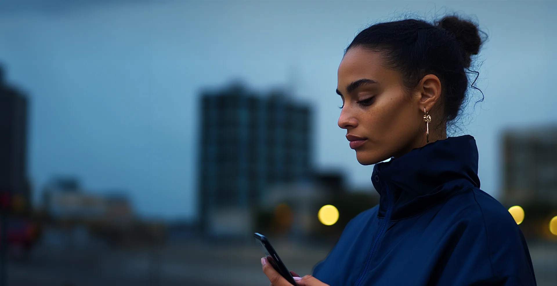 Mulher de perfil, com o cabelo preso e brinco dourado, olhando para o celular ao entardecer, com prédios desfocados ao fundo
