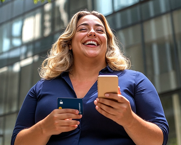 Mulher loira sorrindo, segurando um celular e um cartão de crédito RecargaPay, em frente a prédios