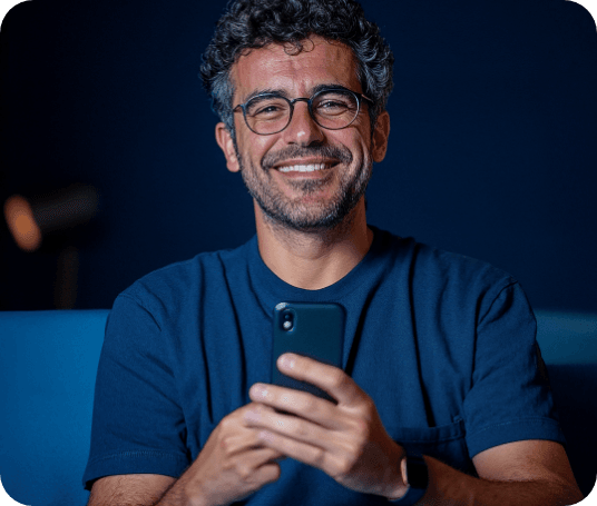 Homem de óculos e barba grisalha, de camiseta azul, sorrindo e segurando um celular
