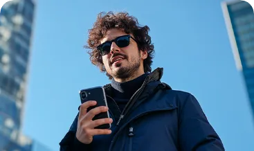 Homem de óculos escuros e cabelo cacheado, de jaqueta azul, segurando um celular, visto de baixo contra prédios e céu azul