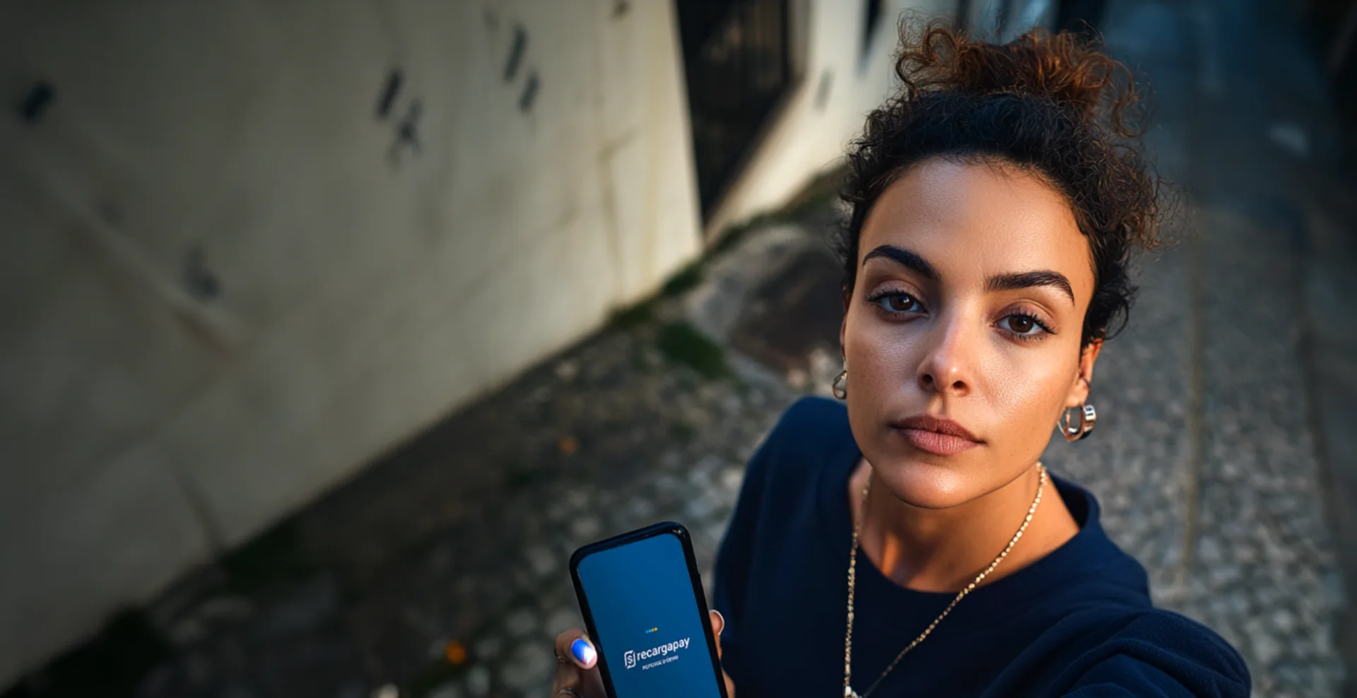 Mulher de coque e colar, de blusa azul-marinho, segurando o celular em uma rua de paralelepípedo, em foto de ângulo alto