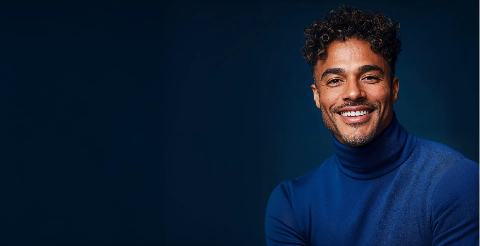 Homem sorridente de cabelo cacheado e blusa de gola alta azul-marinho, em fundo azul-escuro