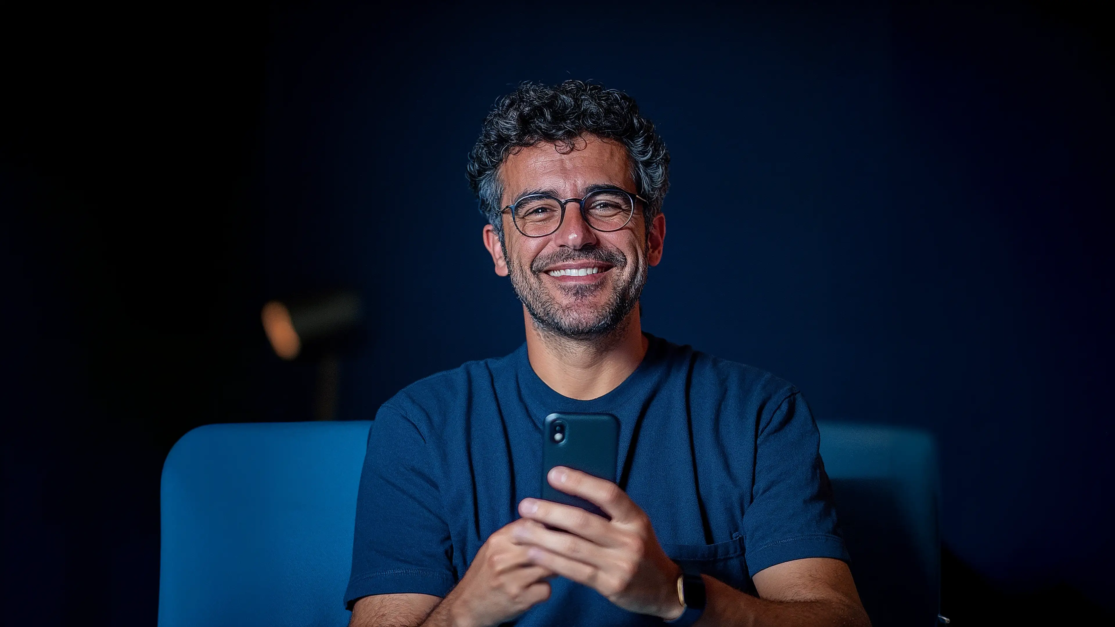 Homem de óculos e cabelo cacheado sorrindo e segurando um celular, em fundo azul-escuro