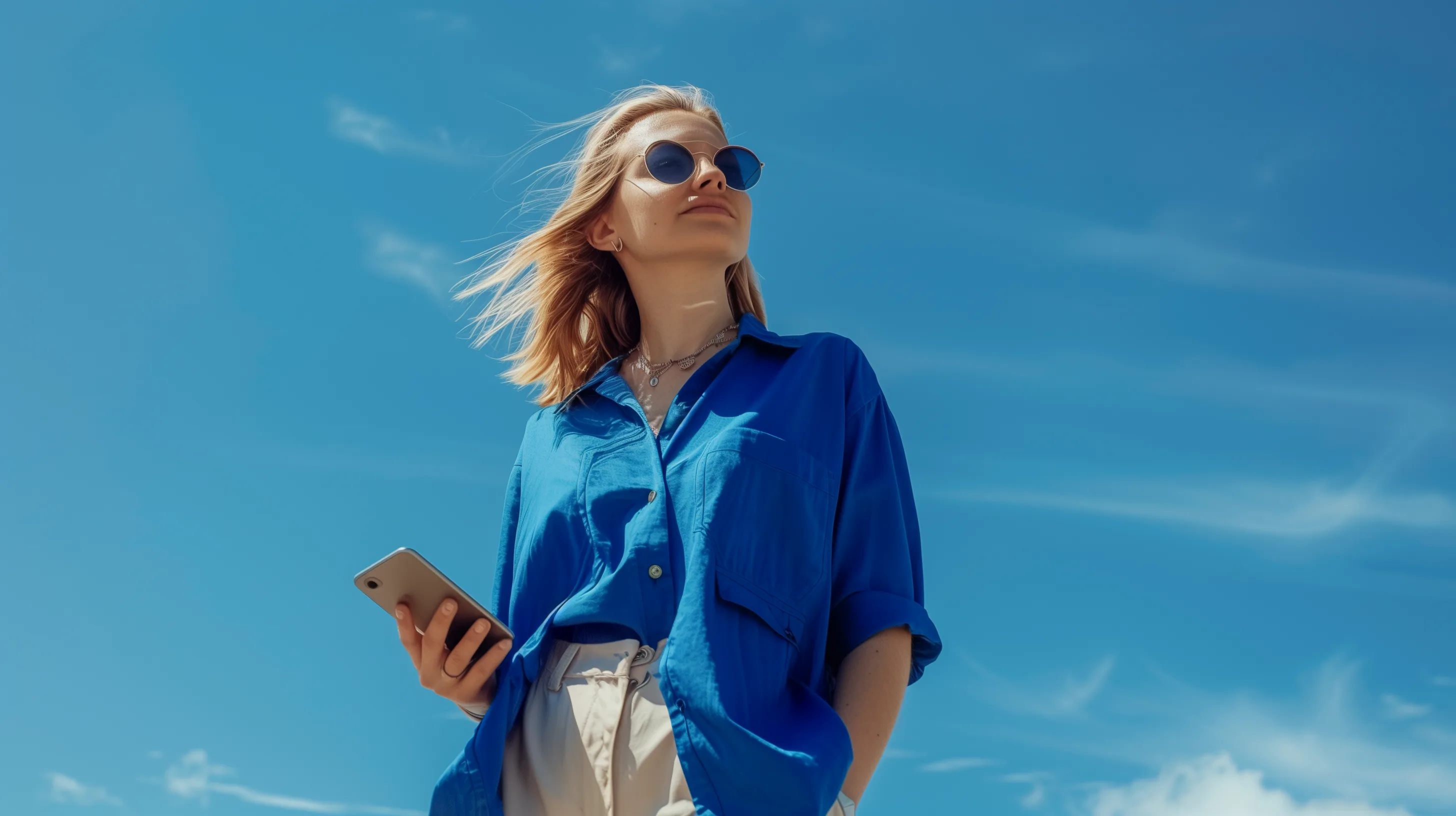 Mulher de camisa azul e óculos de sol usando o celular ao ar livre, com céu azul ao fundo