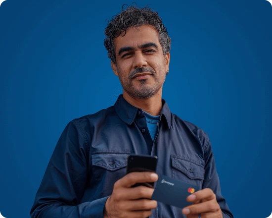 Homem sorridente de cabelos cacheados escuros e barba, vestindo camisa azul marinho, segurando um smartphone com as duas mãos em um gesto de uso do aplicativo.