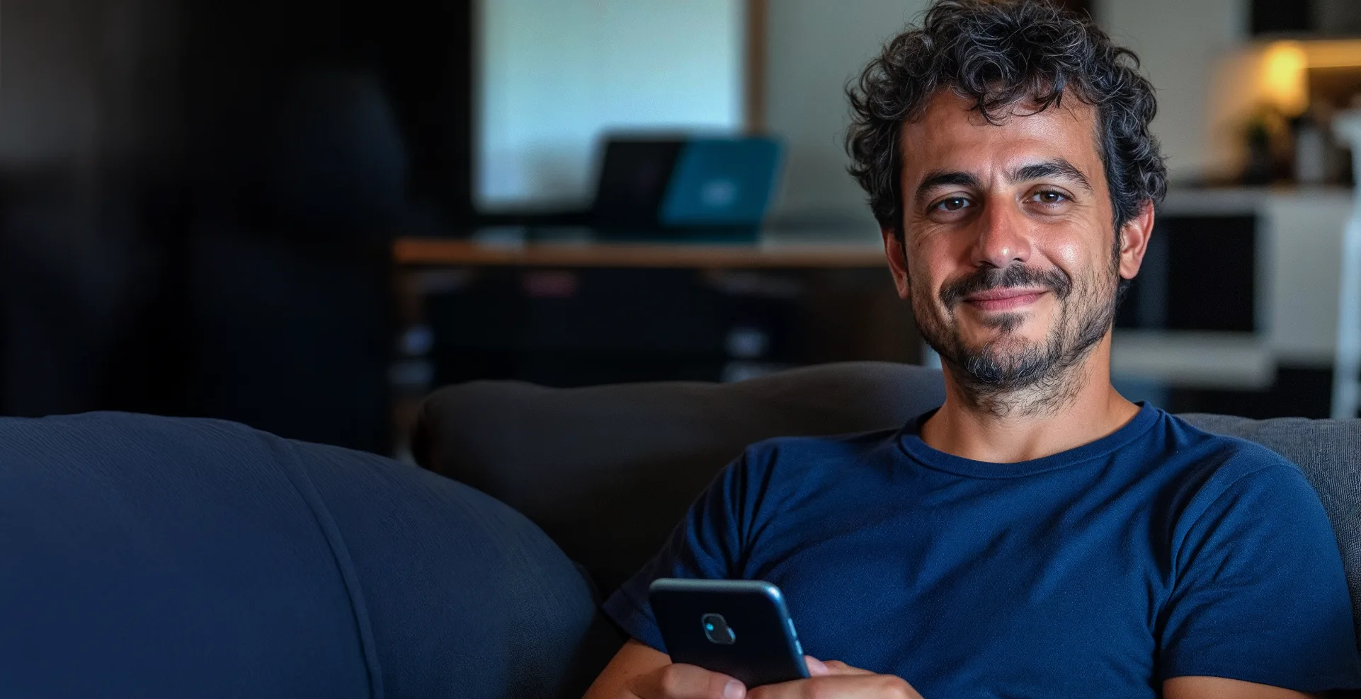 Homem de camiseta azul sorrindo, sentado em um sofá com o celular na mão, em um home office