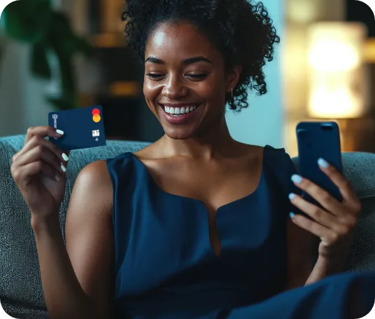 Mulher de cabelo cacheado sorrindo, sentada em um sofá, com um cartão Mastercard em uma mão e o celular na outra
