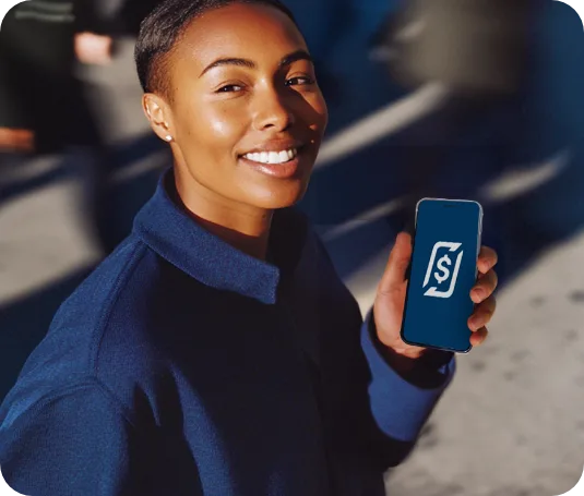Mulher sorridente de cabelo curto, com jaqueta azul, segurando um celular com o app RecargaPay em uma rua movimentada