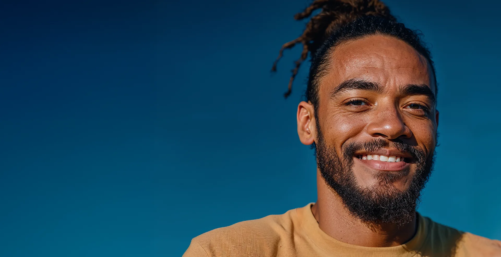 Homem sorrindo, com cabelo preso e barba, segurando um celular contra um céu azul