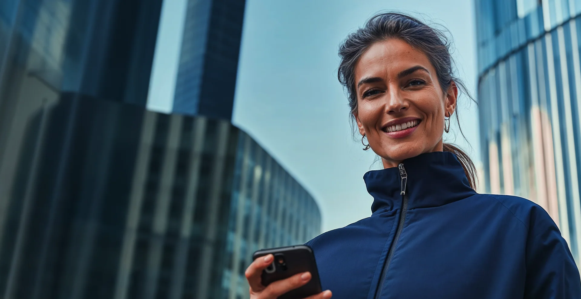 Mulher sorrindo ao usar o celular em frente a prédios.