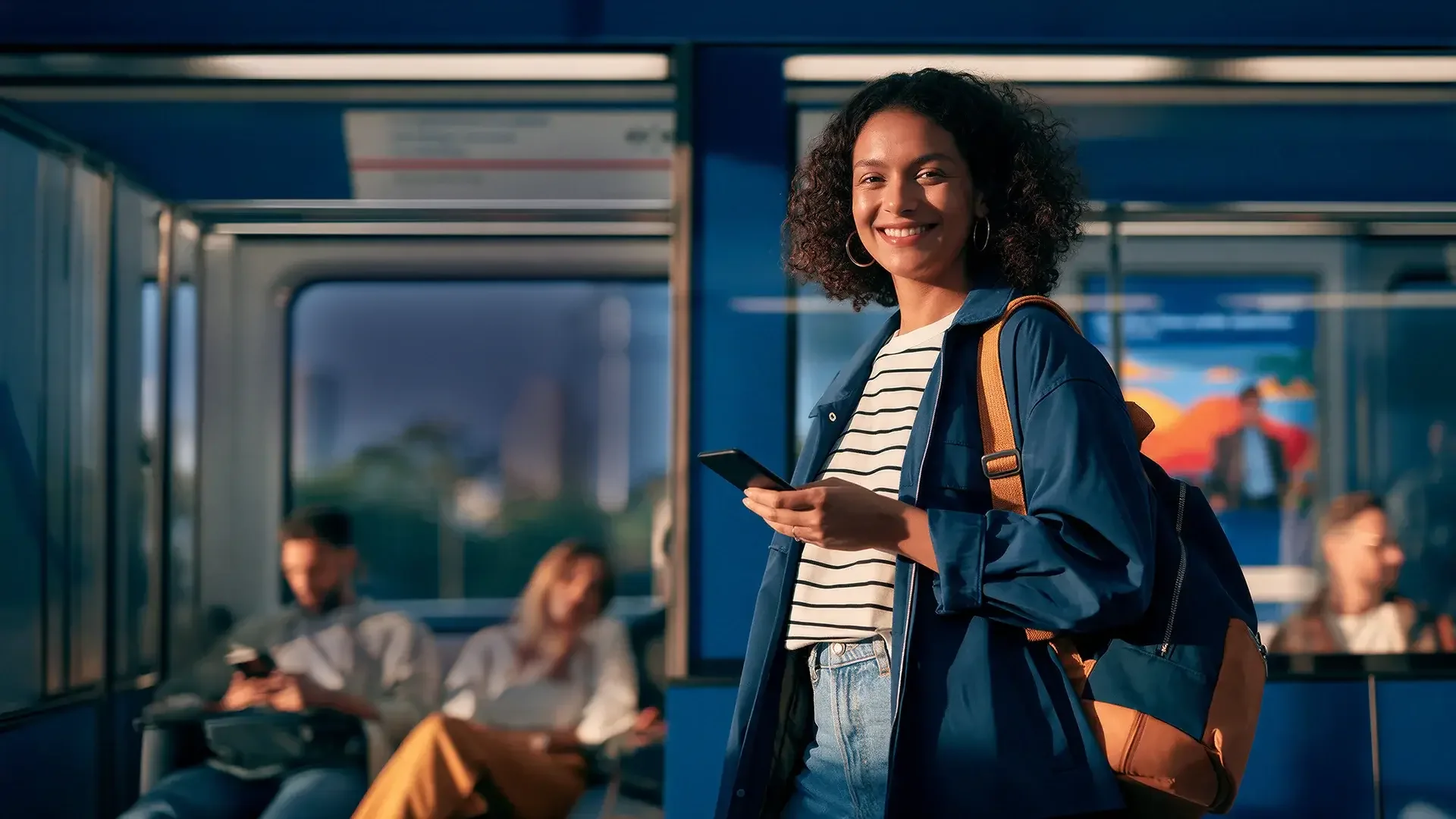 Mulher de cabelo cacheado sorrindo, com mochila e celular na mão, em um vagão de metrô com passageiros ao fundo