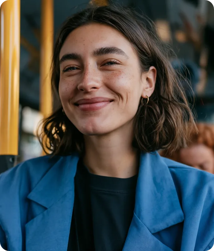 Mulher sorrindo, de casaco azul, sentada em um ônibus com barras amarelas ao fundo