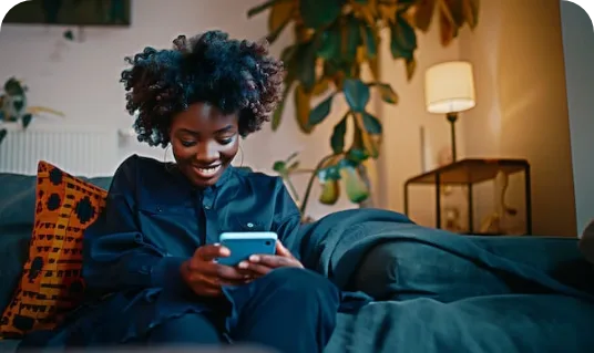 Mulher de cabelo afro sorrindo enquanto olha o celular, sentada em um sofá de casa