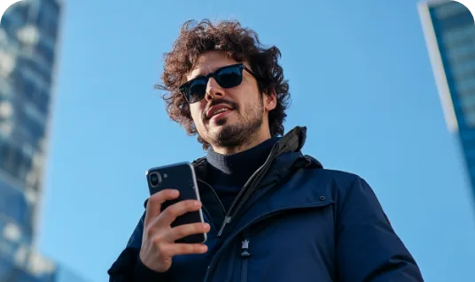 Homem de óculos escuros e casaco azul olhando para o celular, com prédios e céu azul ao fundo
