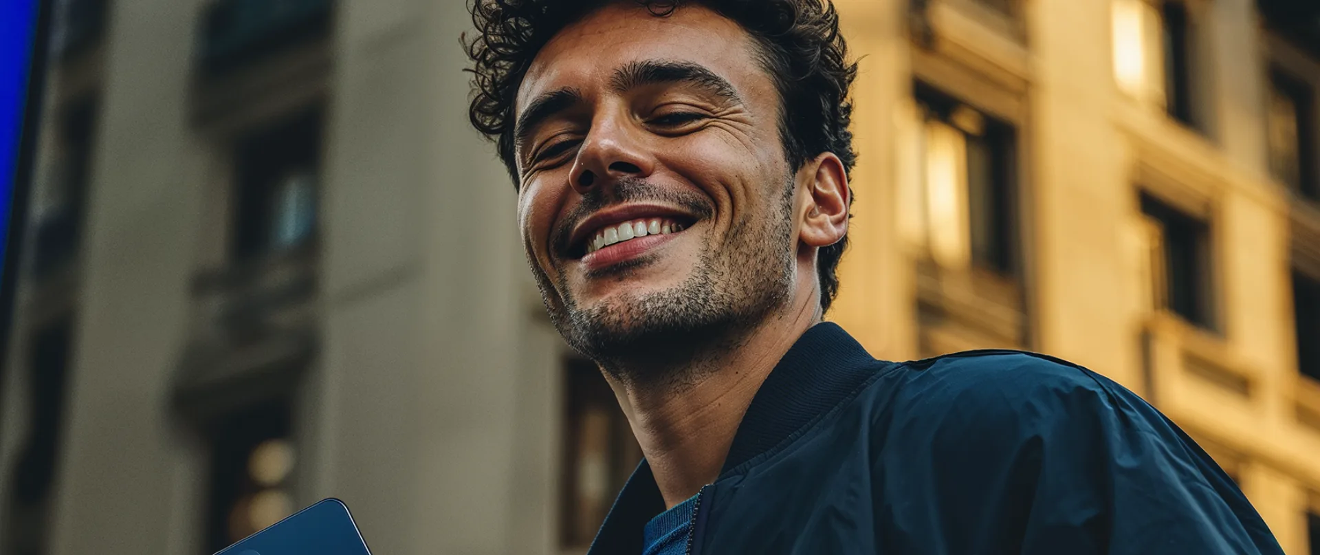 Homem sorrindo olhando para a câmera de baixo para cima, com prédios iluminados ao fundo e o celular na mão