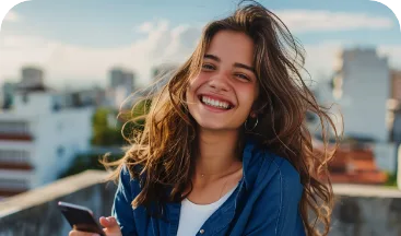 Mulher sorrindo com o cabelo ao vento, segurando um celular em frente a prédios da cidade