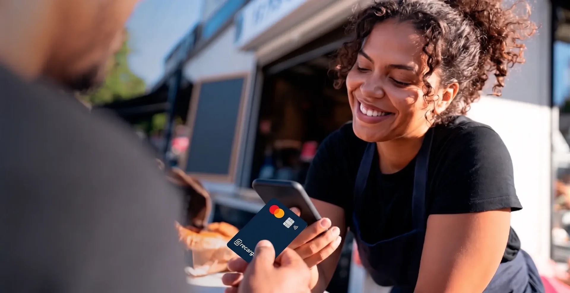Comerciante sorridente de cabelo cacheado recebendo um pagamento por aproximação do cartão no celular, em sua banca de rua