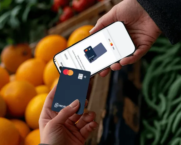 Mãos aproximando um cartão de crédito RecargaPay de um celular, em uma banca de frutas, para receber um pagamento por aproximação com Tap to Pay